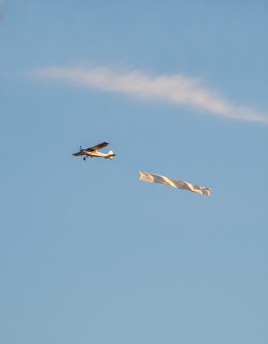A banner-tow plane flying high across a clear blue sky trailing a long fabric banner